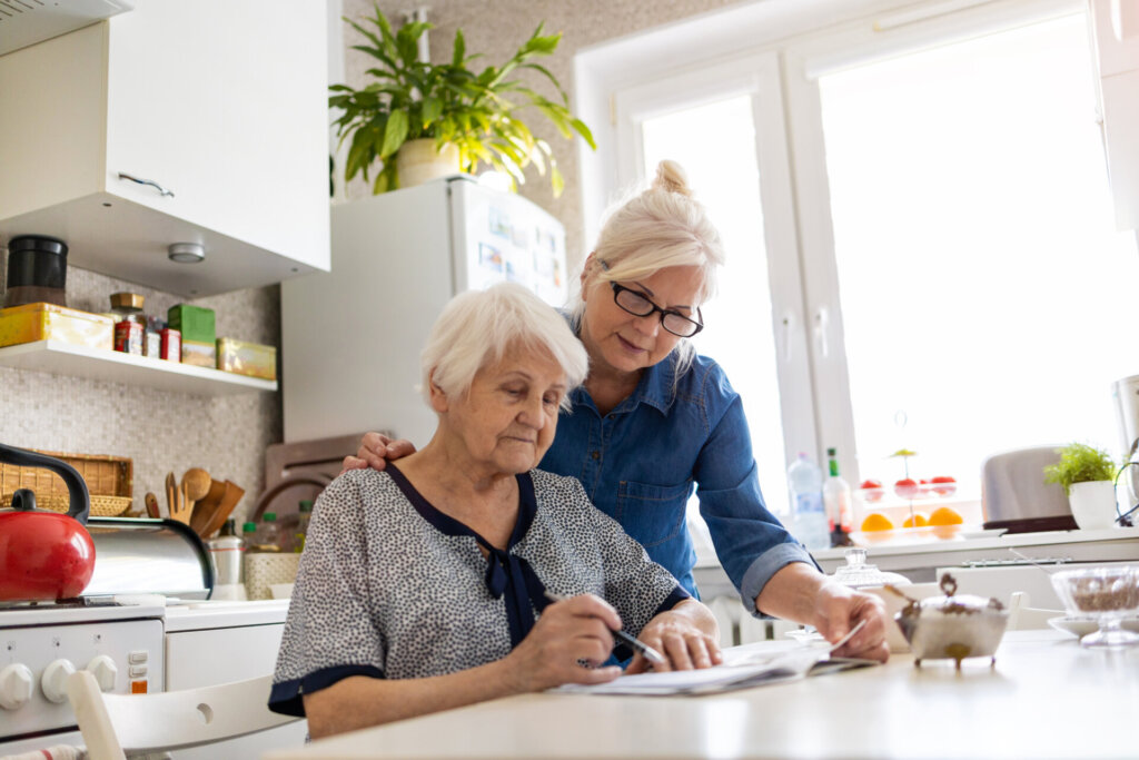 A caregiver looking over an elderly woman’s shoulder. Both looking at paperwork on the kitchen table.